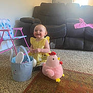Amaya joined the competition — help win amazing prizes! toddler, child, yellow_dress, smile, plush_toy, pink_chicken, blue_basket, bunny_ears, carpet, rug, living_room, sofa, couch, kids_chair, toy, indoor, happy, person, floor, home