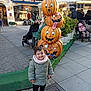 child, toddler, pumpkin, jack_o_lantern, outdoor, market, smiling, jacket, scarf, stroller, people, pavement, plants, decorations, autumn, holiday, festive, funny_faces, public_space, daytime