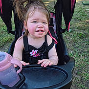 Nevaeh is registered to the contest to win money with this photo: child, toddler, girl, portrait, smiling, outdoor, stroller, baby_bottle, pink_bottle, black_clothing, harness, ponytail, brown_hair, grass, park, seated, playground, daylight, close_up, happy