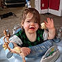 child, toddler, messy_face, food_on_face, exersaucer, toy, carpet, living_room, smiling, waving, brown_hair, green_shirt, playtime, seat, tray, infant, furniture, background_person, bed, blanket