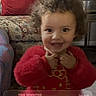 toddler, child, smiling, red_clothing, curly_hair, indoor, armchair, pillow, furniture, phone_screen, lock_screen, notification, time_display, date_display, portrait, person, happy, couch, home, cute