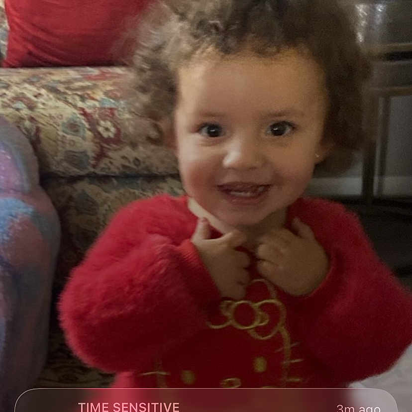 Roselina joined the competition — help win amazing prizes! toddler, child, smiling, red_clothing, curly_hair, indoor, armchair, pillow, furniture, phone_screen, lock_screen, notification, time_display, date_display, portrait, person, happy, couch, home, cute