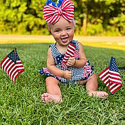 Oaklee is registered to the contest to win money with this photo: baby, child, grass, outdoor, flag, american_flag, headband, bow, smile, happy, patriotic, summer, nature, person, sitting, greenery, cute, celebration, holiday, dress