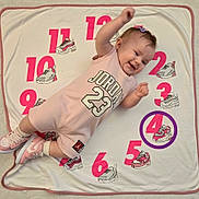 Lilah joined the competition — help win amazing prizes! baby, child, pink_clothing, jordan_23, sneakers, shoes, blanket, numbers, smiling, happy, lying_down, cute, indoor, footwear, hairclip, raising_arm, playful, pattern, purple_circle, white_background