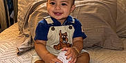 Thiago is registered to the contest to win money with this photo: toddler, child, smiling, bed, pillow, overalls, sneakers, sitting, indoors, bedroom, cute, happy, clothing, footwear, person, head, face, furniture, blanket, decor