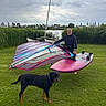 man, dog, grass, windsurfing_board, outdoor, cloudy_sky, water, reeds, greenery, sports_equipment, pet, black_dog, brown_dog, person, leisure, nature, activity, summer, lake, casual