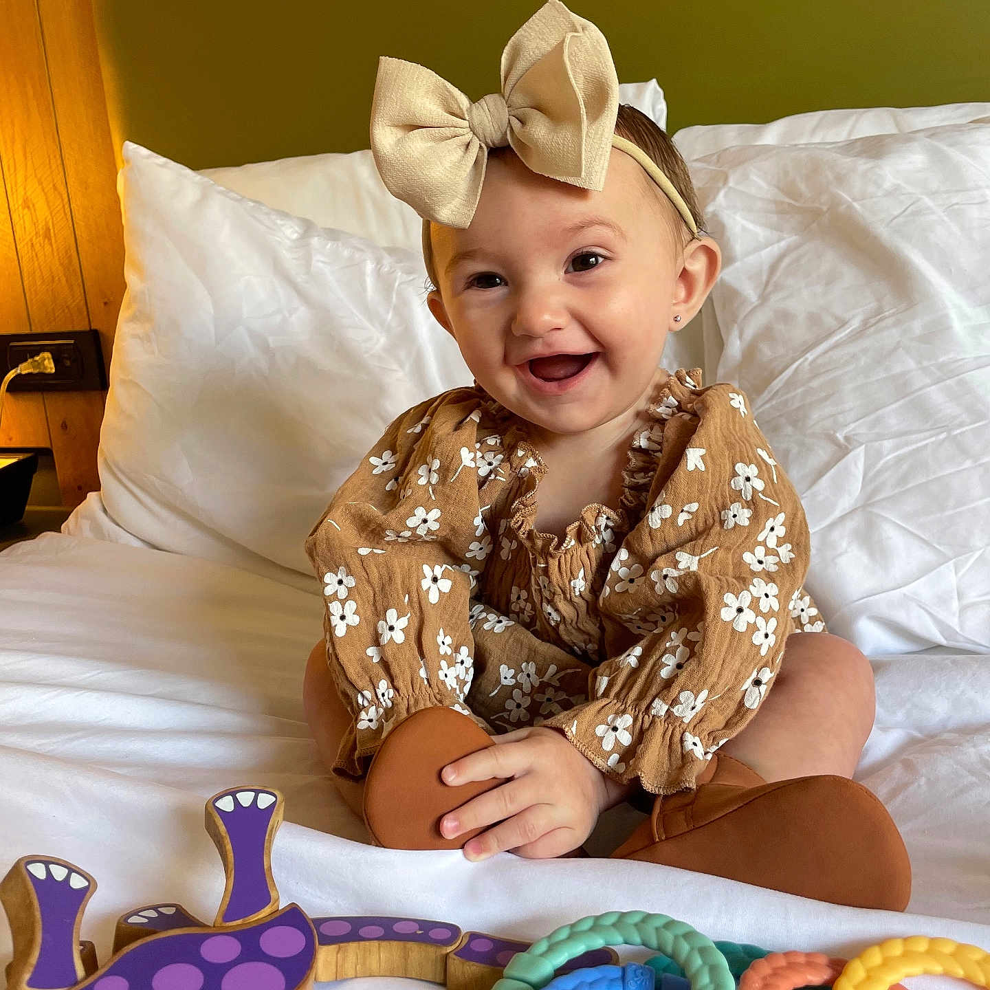 Eliyanah joined the competition — help win amazing prizes! baby, bed, bow, child, colorful_toys, cute, dress, floral_pattern, green_wall, happy, headband, indoors, playful, rings, sitting, smiling, toy, warm_lighting, white_bedding, wooden_toy