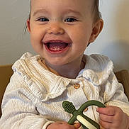 Isla is registered to the contest to win money with this photo: baby, child, smile, teether, toy, white_clothing, happy, face, hands, indoor, portrait, cute, infant, sitting, young_child, closeup, skin, person, mouth_open, eyes
