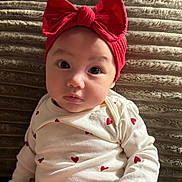 Leilani is registered to the contest to win money with this photo: baby, child, red_bow, headband, white_onesie, heart_pattern, sitting, face, portrait, indoors, cozy, cushion, blanket, cute, infant, looking, expression, soft_lighting, young, adorable