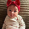 baby, child, red_bow, headband, white_onesie, heart_pattern, sitting, face, portrait, indoors, cozy, cushion, blanket, cute, infant, looking, expression, soft_lighting, young, adorable