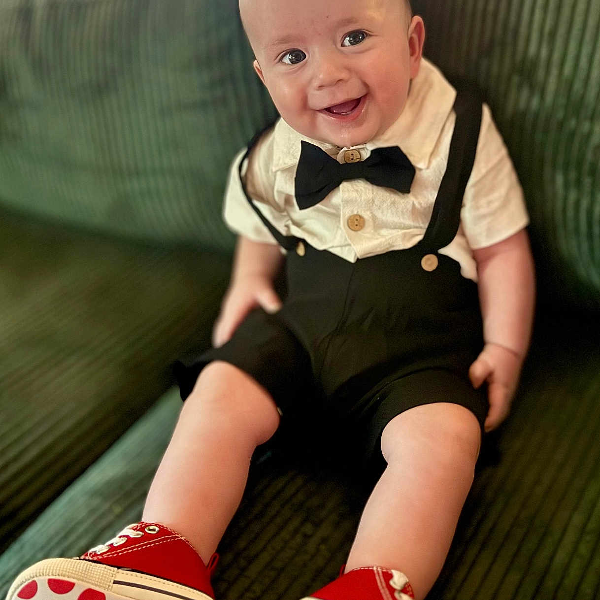 Alden-Paul is registered to the contest to win money with this photo: baby, infant, smiling, bow_tie, suspenders, red_shoes, polka_dot, couch, green_couch, happy, cute, child, indoors, portrait, sitting, clothing, footwear, person, fashion, baby_shoes