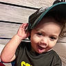 baseball_cap, black_shirt, casual_clothing, child, closeup, curly_hair, cute, expressive_eyes, face, happy, headwear, indoor, person, playful, portrait, smiling, toddler, tongue_out, wooden_background, young_child