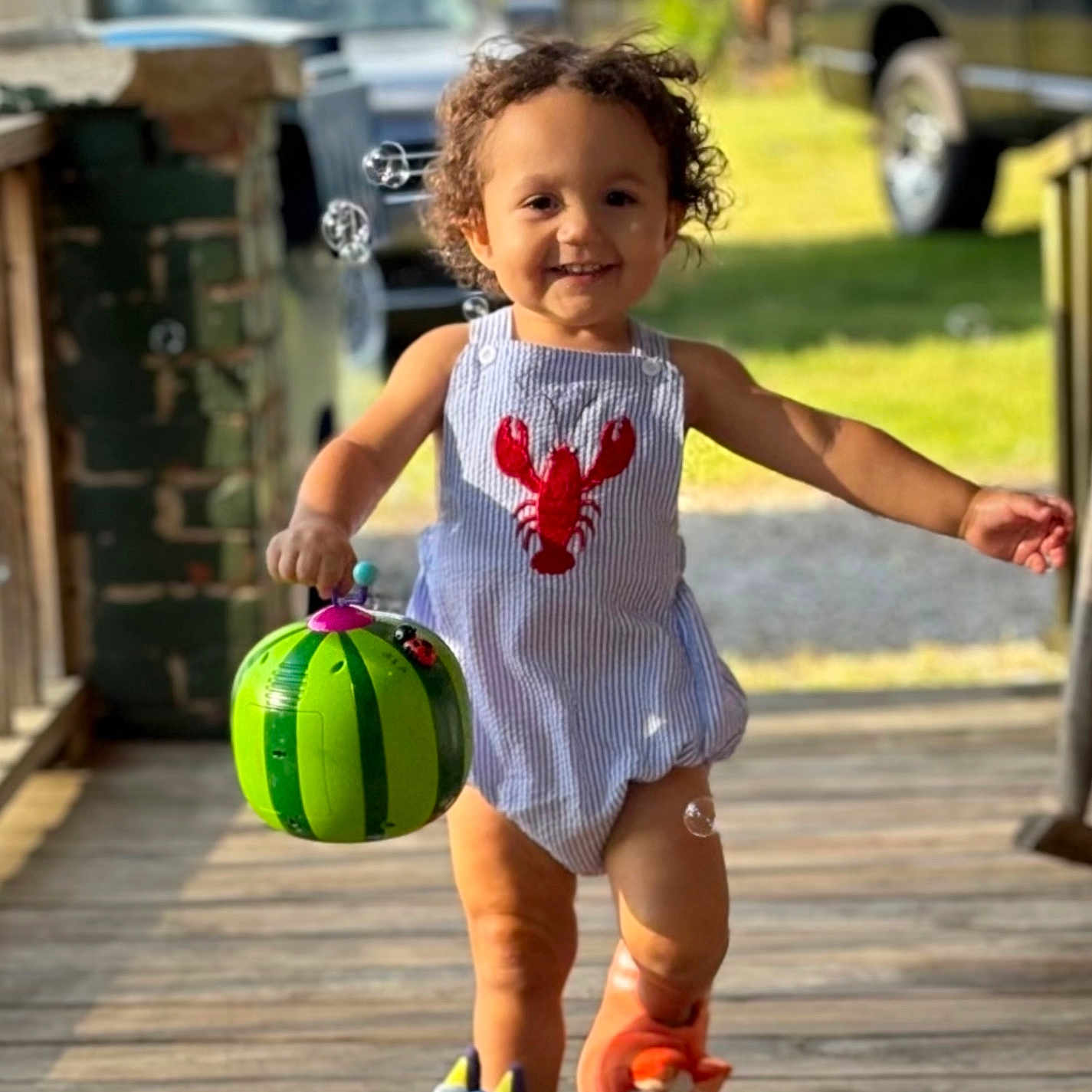 Kashton is registered to the contest to win money with this photo: toddler, child, smiling, outdoor, sunlight, porch, wooden_floor, bubbles, bucket, watermelon, footwear, slippers, clothing, curly_hair, happy, playful, person, daytime, green, background_car