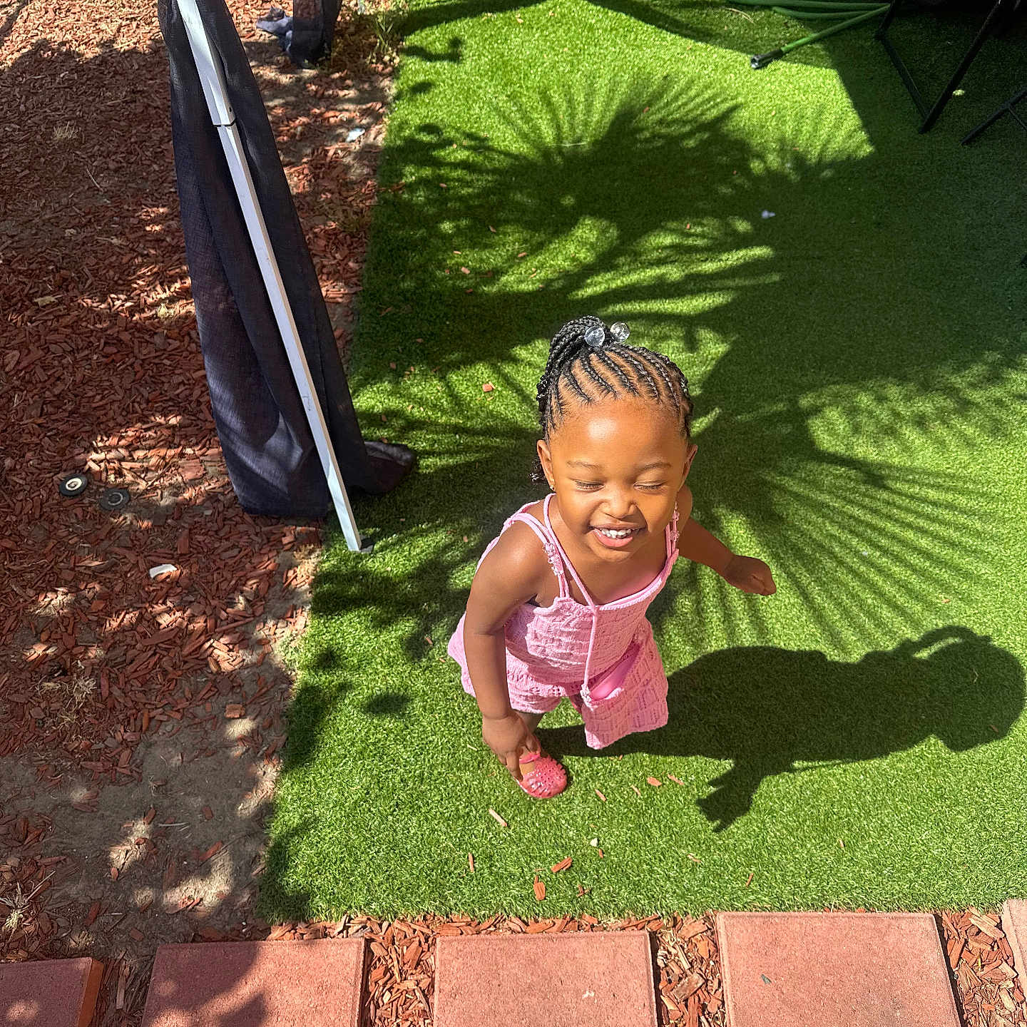Nyleah is registered to the contest to win money with this photo: child, girl, smiling, outdoor, sunlight, shadow, green_grass, braids, pink_dress, happy, playful, pattern, artificial_grass, plant_shadow, footwear, paving_stones, garden, summer, sunny_day, portrait
