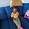 Nyleah joined the competition — help win amazing prizes! child, toddler, smiling, sitting, blue_couch, yellow_shirt, khaki_pants, pink_shoes, hair_braids, golden_pillow, white_blankets, backpack, indoor, happy, person, footwear, cushion, furniture, casual_clothing, relaxed