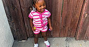 Nyleah joined the competition — help win amazing prizes! child, toddler, smiling, pink_clothing, striped_outfit, braided_hair, beads, wooden_fence, concrete, outdoor, sidewalk, shoes, playful, happy, person, casual_wear, daylight, standing, holding_toys, urban