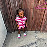 child, toddler, smiling, pink_clothing, striped_outfit, braided_hair, beads, wooden_fence, concrete, outdoor, sidewalk, shoes, playful, happy, person, casual_wear, daylight, standing, holding_toys, urban