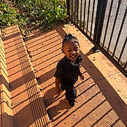Nyleah joined the competition — help win amazing prizes! child, toddler, smiling, outdoor, sunlight, shadow, fence, black_clothing, happy, walking, concrete, plants, greenery, daylight, person, young_child, pattern, playful, shadows, hair_bun