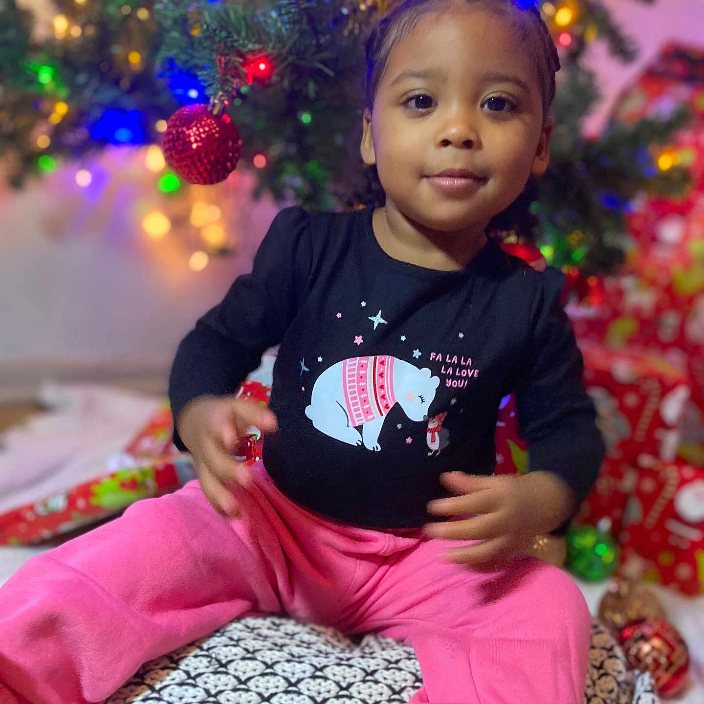 Newlianys is registered to the contest to win money with this photo: black_shirt, braided_hair, child, christmas_tree, cozy, cushion, decorations, face, festive, floor, gift_wrap, holiday, indoor, lights, ornament, pink_pants, polar_bear, portrait, smile, toddler