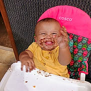 Jonah joined the competition — help win amazing prizes! baby, child, high_chair, chocolate, messy, smiling, happy, yellow_shirt, face, hand, food, indoor, seat, colorful, pattern, playful, person, young_child, dirt, fun