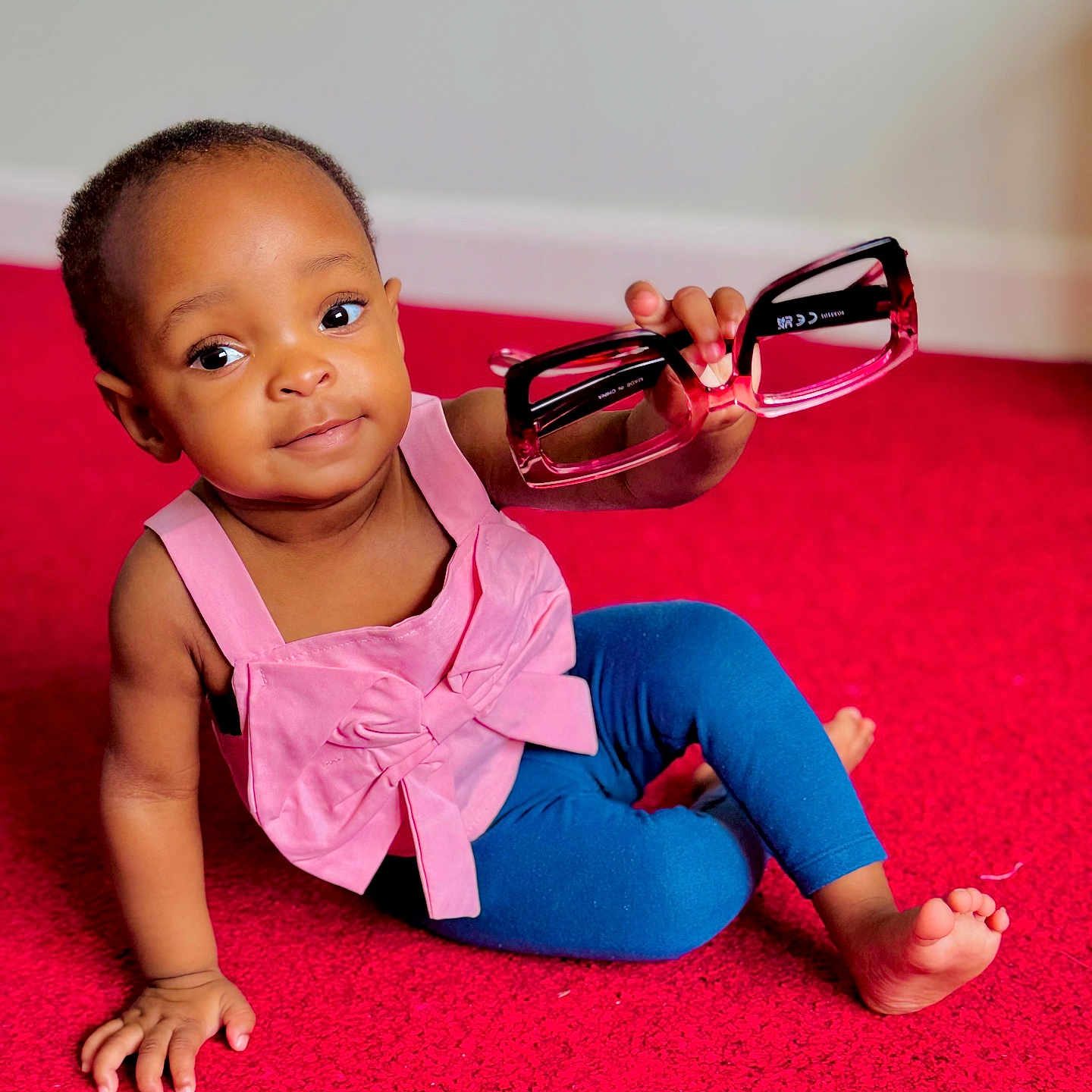 Kiley is registered to the contest to win money with this photo: baby, blue_leggings, child, cute, expression, floor, foot, glasses, hand, holding, indoor, person, pink_top, playing, portrait, red_carpet, sitting, smiling, toddler, young_child