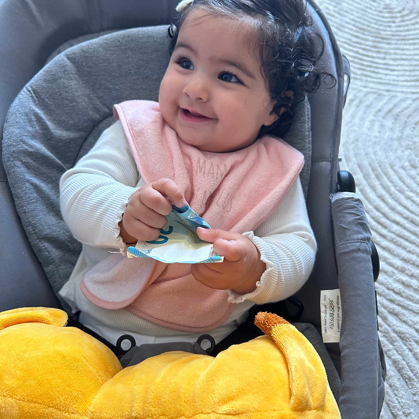 Issra participe au concours pour gagner de l'argent avec cette photo : baby, baby_seat, carpet, child, curly_hair, cushion, cute, happy, holding, indoor, infant, person, pink_bib, plush_toy, portrait, seated, smiling, soft_toys, white_clothing, yellow_toy