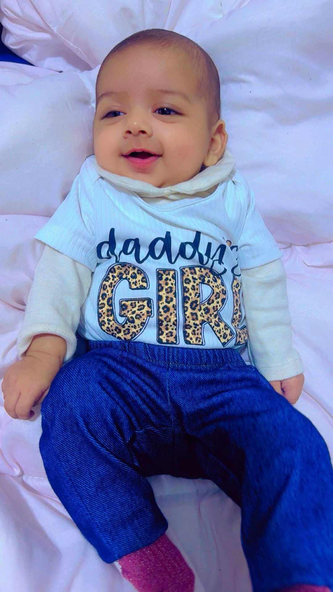 Amreen is registered to the contest to win money with this photo: baby, child, smiling, cute, infant, clothing, shirt, pants, socks, bed, blanket, portrait, lying_down, happy, face, young_child, fabric, text_on_clothing, indoor, person