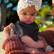 Ellie Grace is registered to the contest to win money with this photo: baby, child, white_bow, headwear, black_shirt, adult_hand, knit_sweater, outdoor, sunlight, greenery, soft_focus, portrait, cute, infant, person, skin, face, holding, warm_colors, closeup