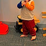 Fischer joined the competition — help win amazing prizes! baby, barefoot, blond_hair, blue_sleeve, carpet, child, curious_expression, face, holding_object, indoor, long_sleeve_shirt, orange_sleeve, person, playroom, red_pants, standing, toddler, toy, toy_truck, wall