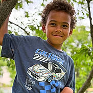 Enzo a rejoint le concours — aidez-le/la à gagner de superbes lots ! child, boy, curly_hair, tree, branch, tshirt, racing_car_graphic, outdoor, greenery, nature, smiling, portrait, casual_clothing, daylight, young, playful, happy, leaves, background_blur, person