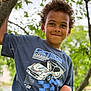 child, boy, curly_hair, tree, branch, tshirt, racing_car_graphic, outdoor, greenery, nature, smiling, portrait, casual_clothing, daylight, young, playful, happy, leaves, background_blur, person