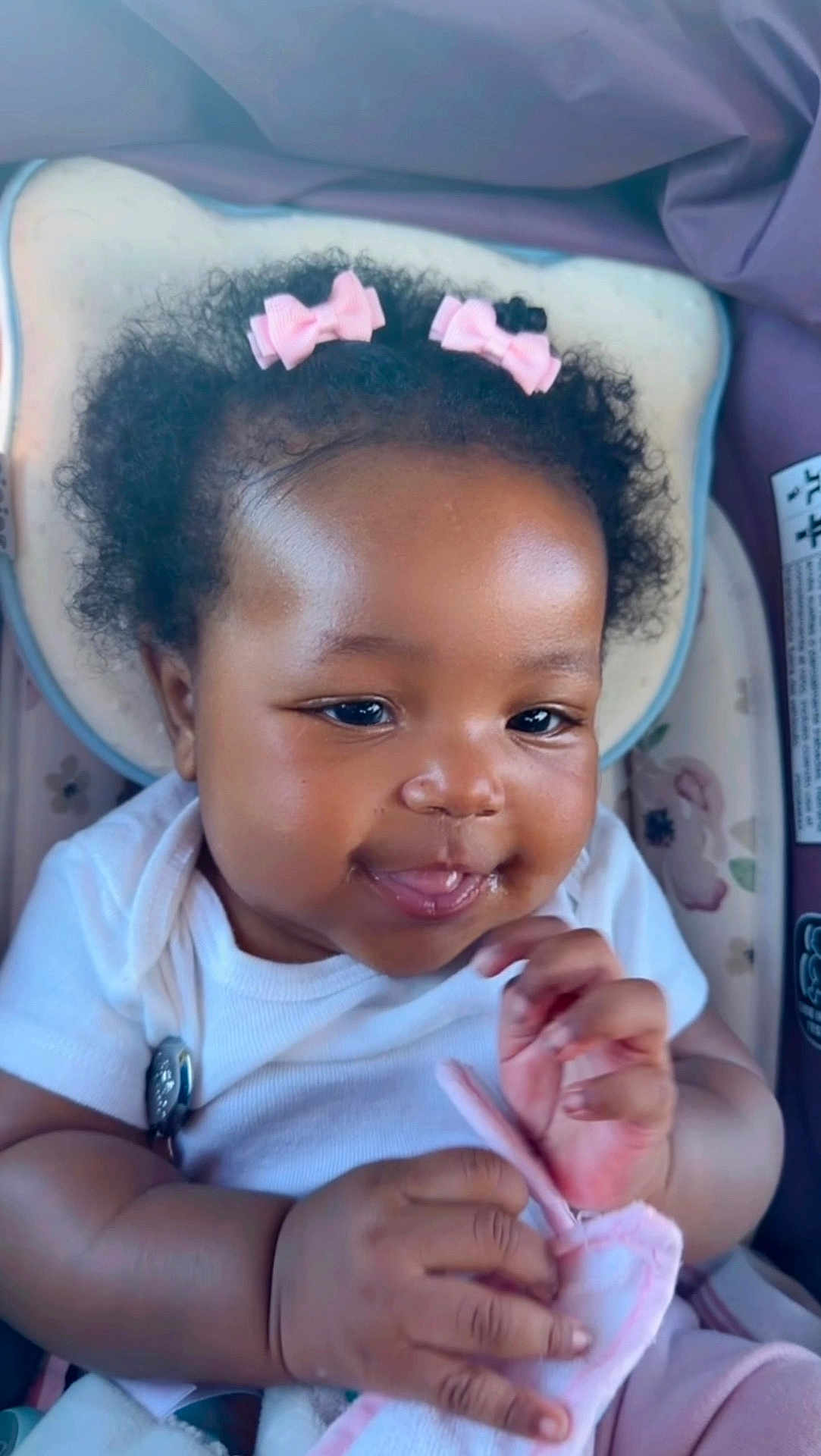 Skylar is registered to the contest to win money with this photo: baby, infant, curly_hair, pink_bows, hair_bows, onesie, white_clothing, car_seat, headrest, chubby_cheeks, smile, smiling, hands, fingers, soft_toy, blanket, clip, close_up, portrait, adorable