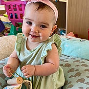 Sierra is registered to the contest to win money with this photo: baby, child, infant, smiling, headband, pink, dress, green, toy, book, fabric, elephant_pattern, cushion, indoor, cute, happy, small_hands, face, person, play