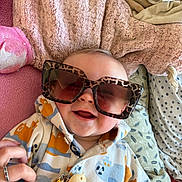 Jules participe au concours pour gagner de l'argent avec cette photo : baby, sunglasses, smile, blanket, onesie, hand, patterned_clothing, cheerful, infant, cozy, lying_down, cute, playful, leopard_print, accessory, indoor, happy, child, soft_texture, person