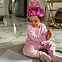 child, toddler, crown, pink_crown, markers, crayons, paper, tile_floor, marble_floor, kitchen, refrigerator, staircase, basket, stuffed_toy, pajamas, curly_hair, sitting, playtime, smiling, indoor