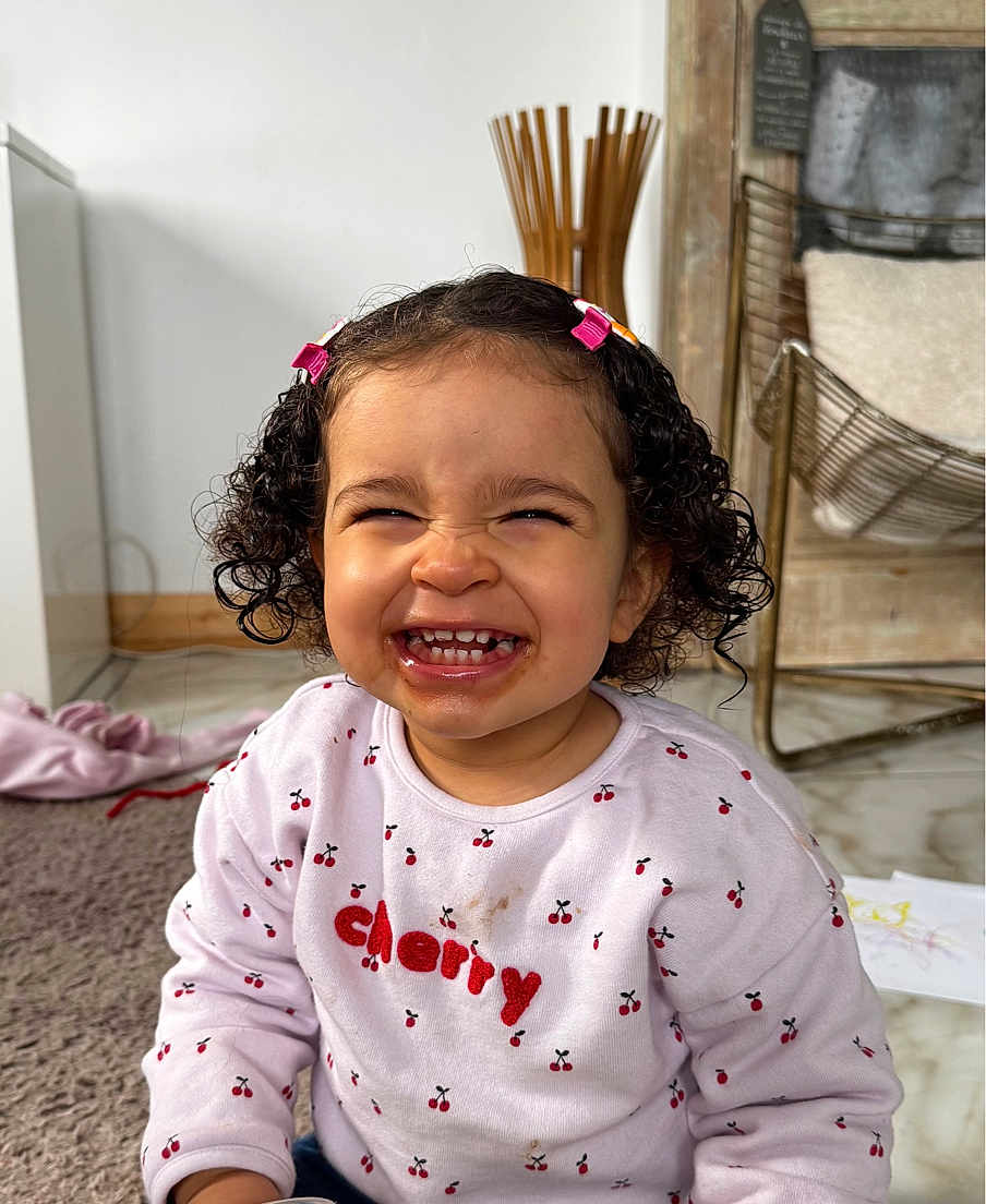 Alya participe au concours pour gagner de l'argent avec cette photo : child, toddler, smiling, grin, curly_hair, hair_clips, sweatshirt, cherry_pattern, stained_clothes, teeth, indoor, carpet, wooden_decor, wicker_chair, playtime, happy_expression, portrait, close_up, playful, messy_face