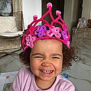 Alya participe au concours pour gagner de l'argent avec cette photo : child, toddler, smile, crown, pink_crown, curly_hair, teeth, pajamas, indoor, marble_floor, drawing_paper, chair, backpack, happy, portrait, closeup, home_interior, playtime, cozy, cute