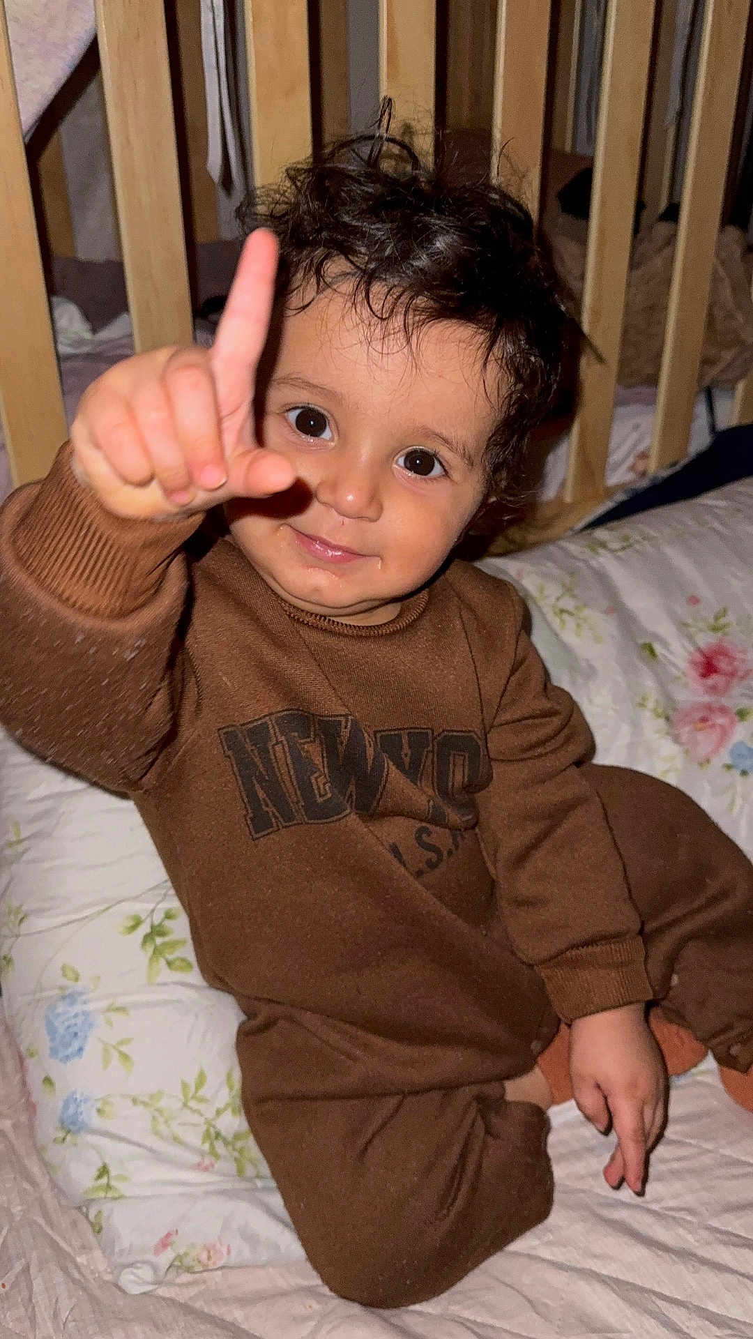 Amir is registered to the contest to win money with this photo: toddler, child, baby, brown_clothing, pointing_finger, curly_hair, big_eyes, crib, pillow, floral_pattern, smile, indoor, cozy, cute, sitting, bed, wooden_bars, playful, portrait, person