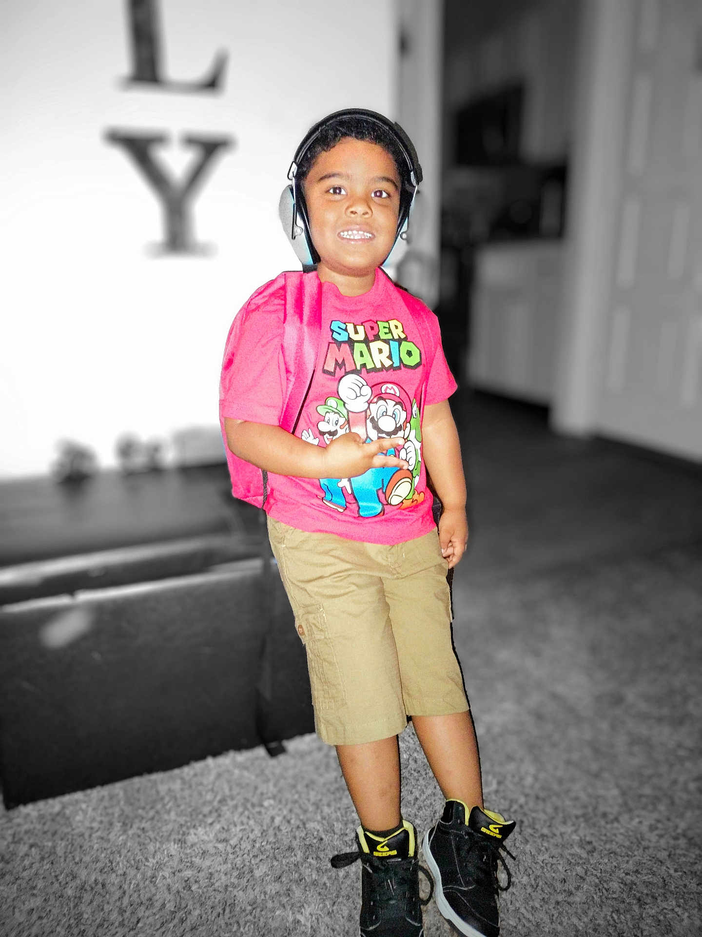 Elijah is registered to the contest to win money with this photo: child, boy, headphones, backpack, super_mario, tshirt, shorts, sneakers, indoor, carpet, furniture, blurred_background, smiling, standing, person, casual_clothing, young, playful, portrait, colorful