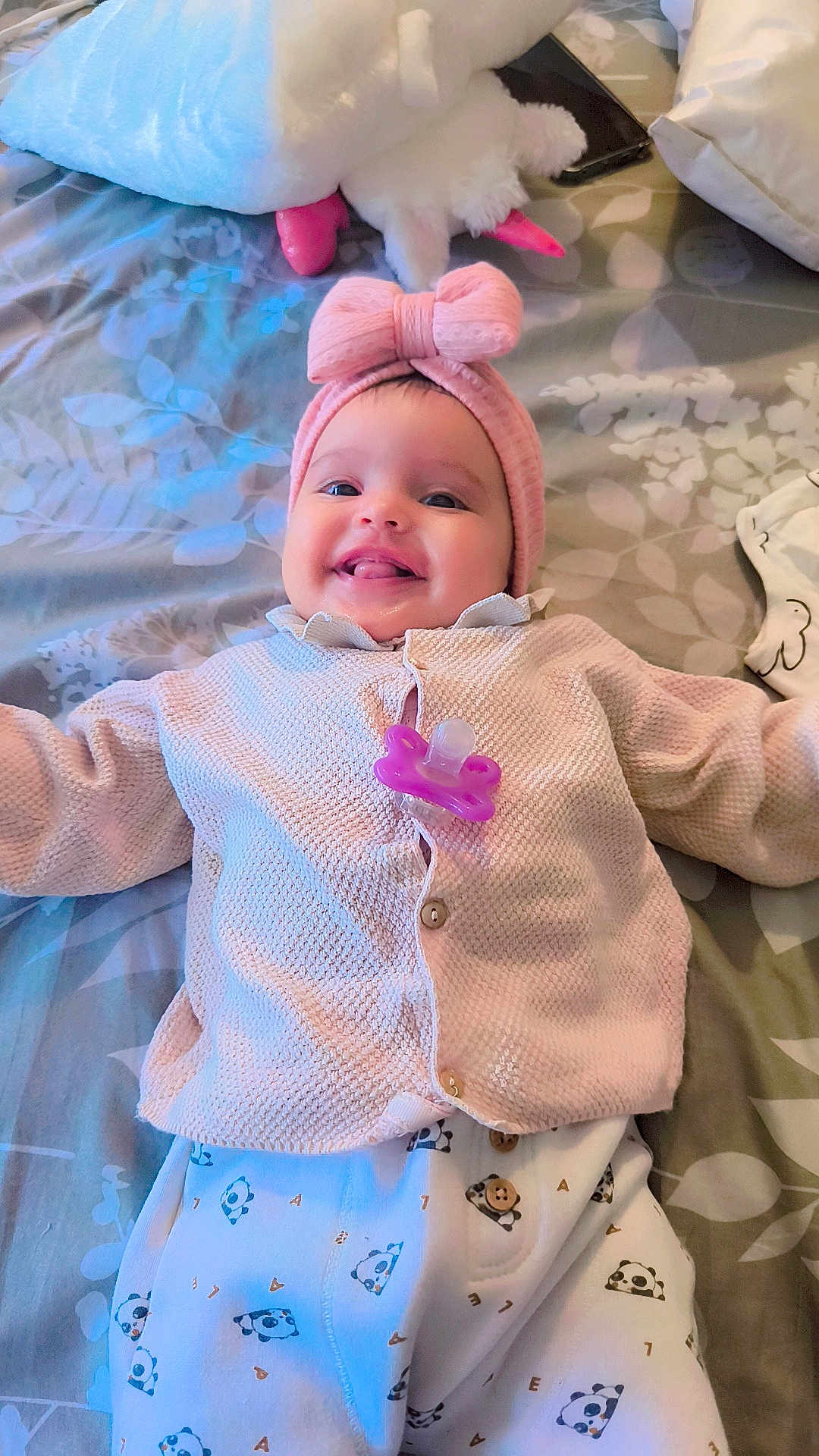 Samia participe au concours pour gagner de l'argent avec cette photo : baby, infant, smiling, pink_bow, headband, pacifier, sweater, pajamas, bed, stuffed_animal, toy, lying_down, happy, cute, child, face, tongue_out, indoor, patterned_bedspread, cozy
