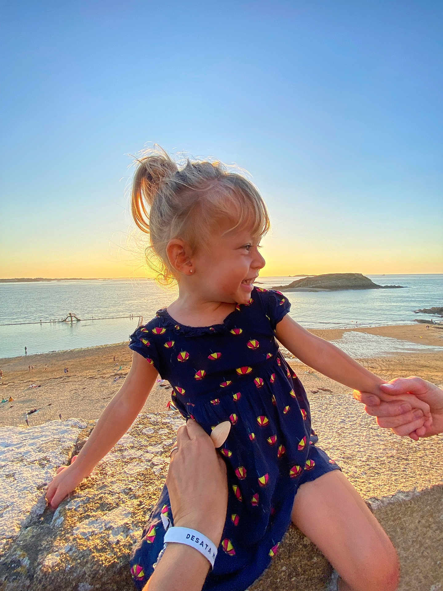 Luciana participe au concours pour gagner de l'argent avec cette photo : child, girl, smiling, sunset, beach, ocean, rock, holding_hands, blue_dress, ponytail, outdoor, water, sand, sky, nature, happy, person, sunlight, vacation, holiday