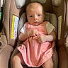 baby, infant, car_seat, pink_dress, white_shoes, socks, child, cute, seated, indoors, looking_at_camera, soft_lighting, car_seat_padding, baby_clothing, small_hands, head_support, baby_face, footwear, baby_girl, comfort