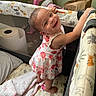 Elizabeth is registered to the contest to win money with this photo: toddler, child, smiling, playpen, floral_dress, indoors, baby_bottle, pillow, blanket, toy, floor, face, hand, foot, room, wall, paper_bag, plastic_packaging, curiosity, happy
