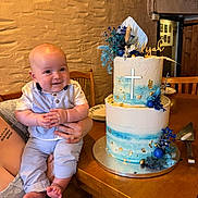 Elijah is registered to the contest to win money with this photo: baby, child, infant, cake, birthday_cake, celebration, blue, white, cross, decoration, table, wooden_furniture, smile, holding, person, indoor, plate, flower_decoration, hand, tattoo