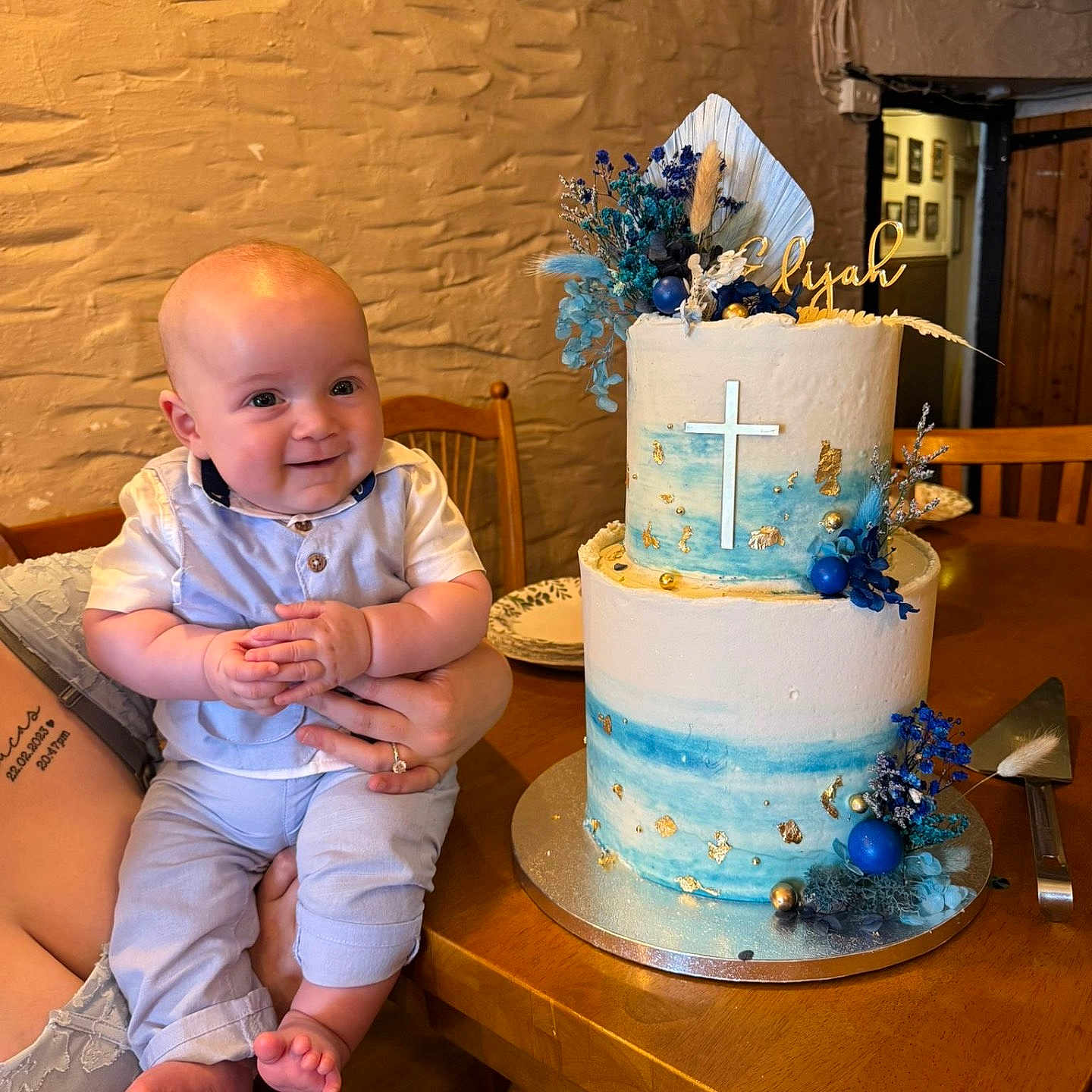 Elijah is registered to the contest to win money with this photo: baby, birthday_cake, blue, cake, celebration, child, cross, decoration, flower_decoration, hand, holding, indoor, infant, person, plate, smile, table, tattoo, white, wooden_furniture