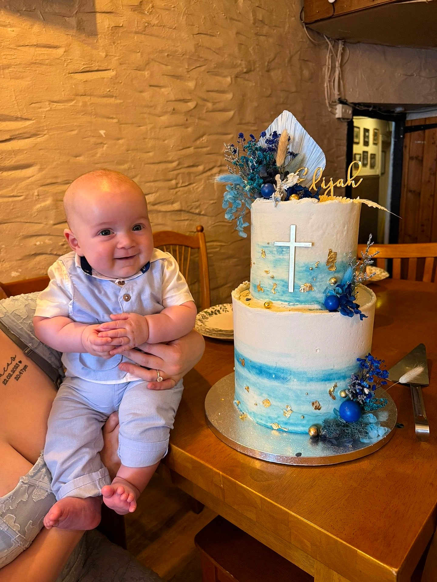 Elijah is registered to the contest to win money with this photo: baby, child, infant, cake, birthday_cake, celebration, blue, white, cross, decoration, table, wooden_furniture, smile, holding, person, indoor, plate, flower_decoration, hand, tattoo