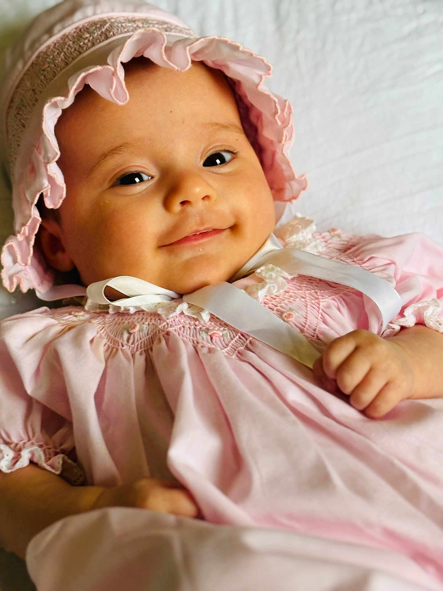 Sophia is registered to the contest to win money with this photo: baby, infant, pink_dress, bonnet, smile, face, child, cute, portrait, laying_down, white_background, soft_lighting, closeup, hands, adorable, young_child, happy, clothing, baby_girl, cozy