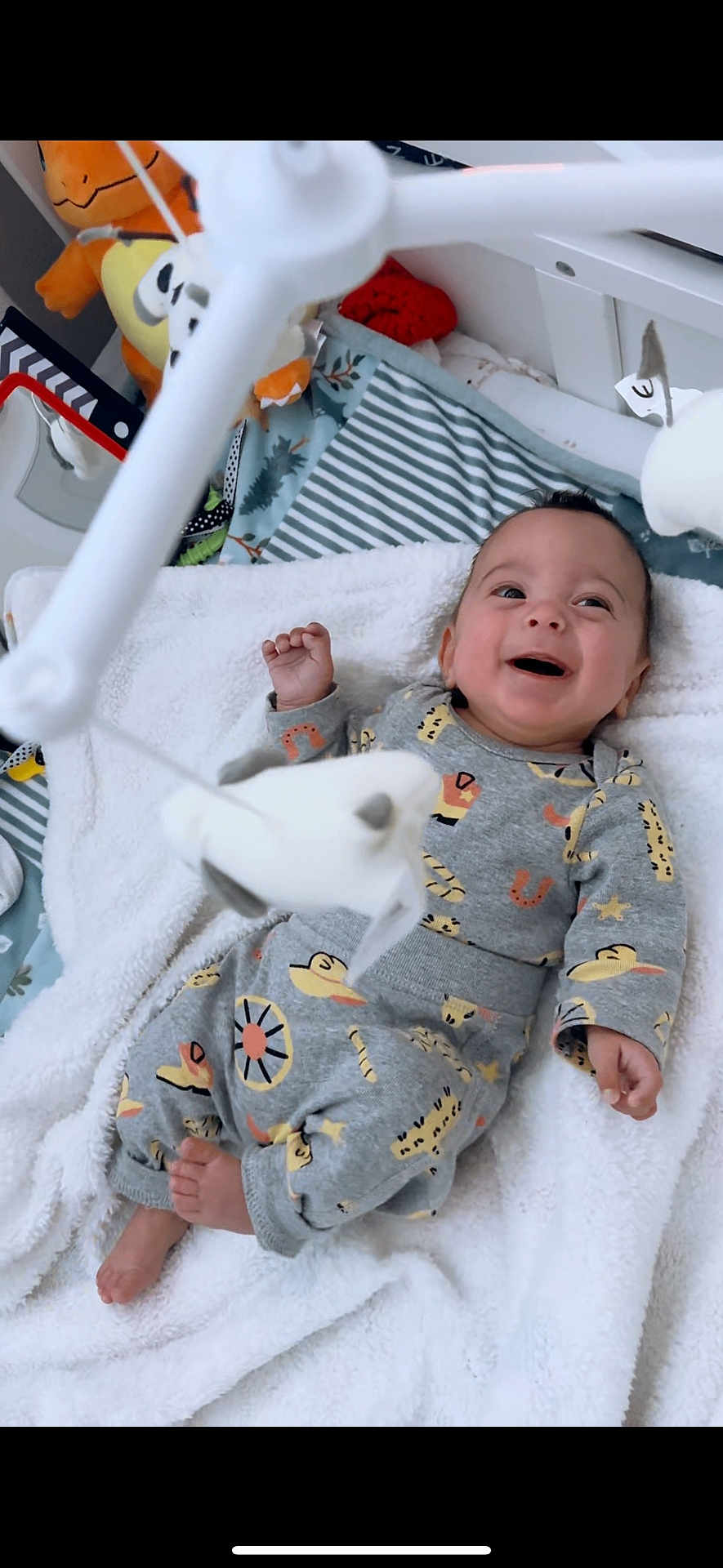 John is registered to the contest to win money with this photo: baby, infant, smiling, blanket, clothing, mobile, toy, indoor, happy, child, cute, patterned_clothes, feet, hand, face, lying_down, soft_toy, nursery, cozy, playtime