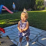 backyard, blue_sky, child, flower, grass, grass_lawn, hedge, house, outdoor, patio, potted_plant, purple_clothing, shadow, slide, smiling, sunlight, toddler, toy_car, trampoline, white_shoes