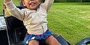 Kaylani is registered to the contest to win money with this photo: arms_raised, black_shoes, child, grass, greenery, hair_bows, happy, jacket, lawnmower, nature, outdoor, park, person, playful, seat, skirt, smiling, sunlight, toddler, tree